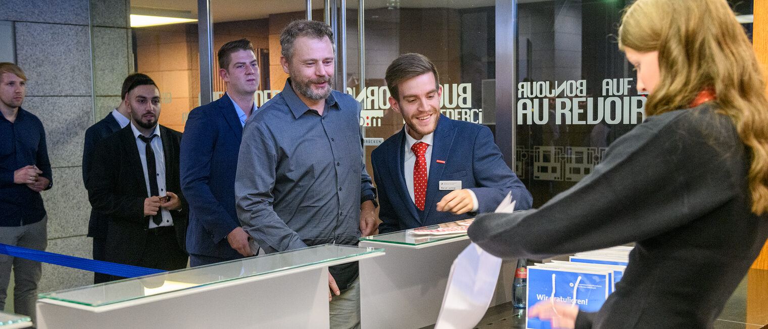 Personen stehen an um ihren Meisterbrief an einem Counter überreicht zu bekommen