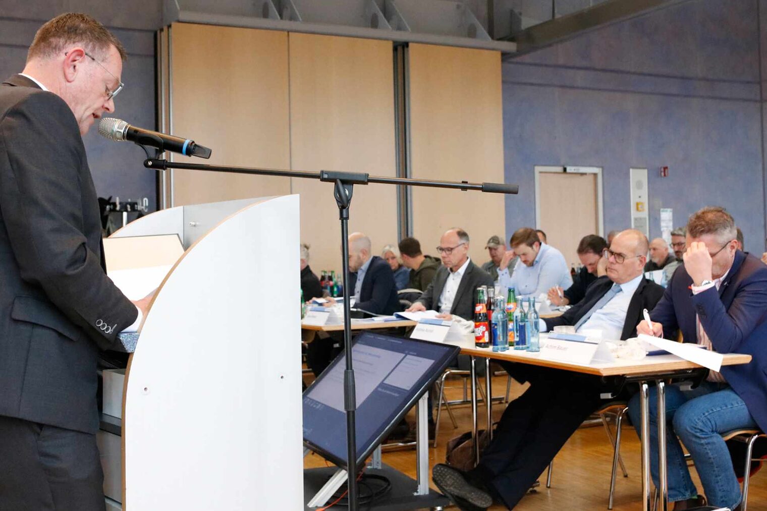 Mann steht links vor Podest und hält eine Rede vor einem Publikum. Man sieht links das Publikum auf Stühlen sitzen. 