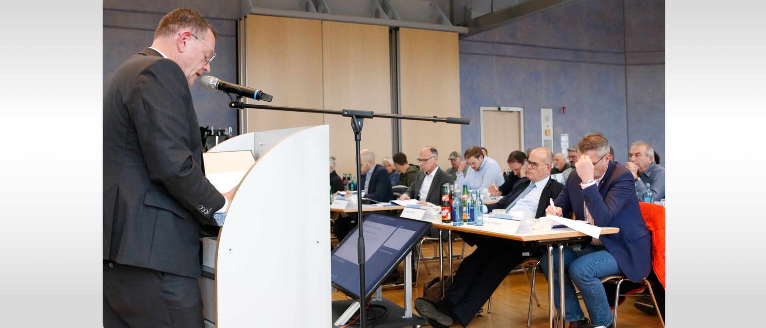 Mann steht links vor Podest und hält eine Rede vor einem Publikum. Man sieht links das Publikum auf Stühlen sitzen. 