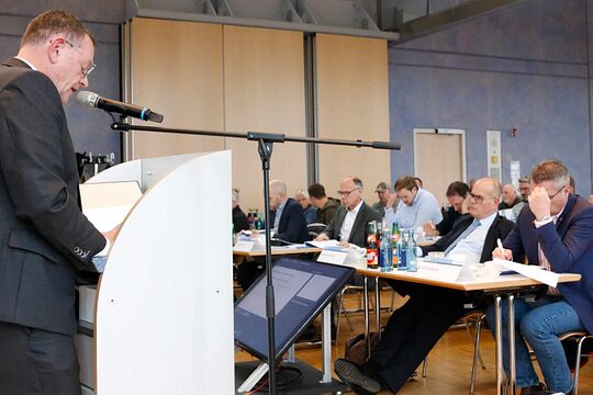 Mann steht links vor Podest und hält eine Rede vor einem Publikum. Man sieht links das Publikum auf Stühlen sitzen. 