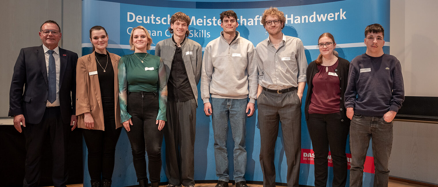 Eine Gruppe junger Menschen steht vor einem Plakat mit dem Titel "Deutsche Meisterschaft im Handwerk" und schaut in die Kamera.