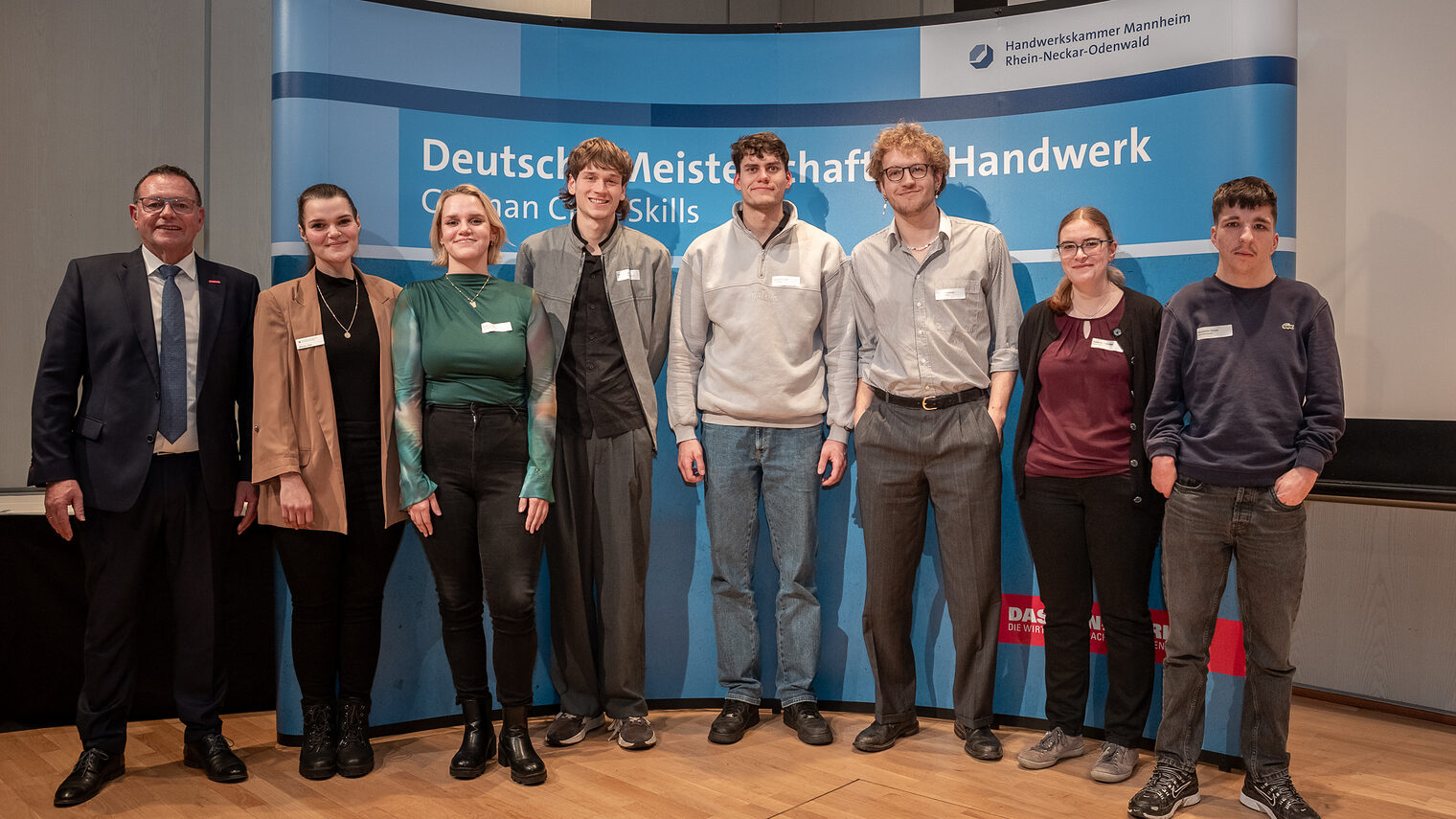 Eine Gruppe junger Menschen steht vor einem Plakat mit dem Titel "Deutsche Meisterschaft im Handwerk" und schaut in die Kamera.