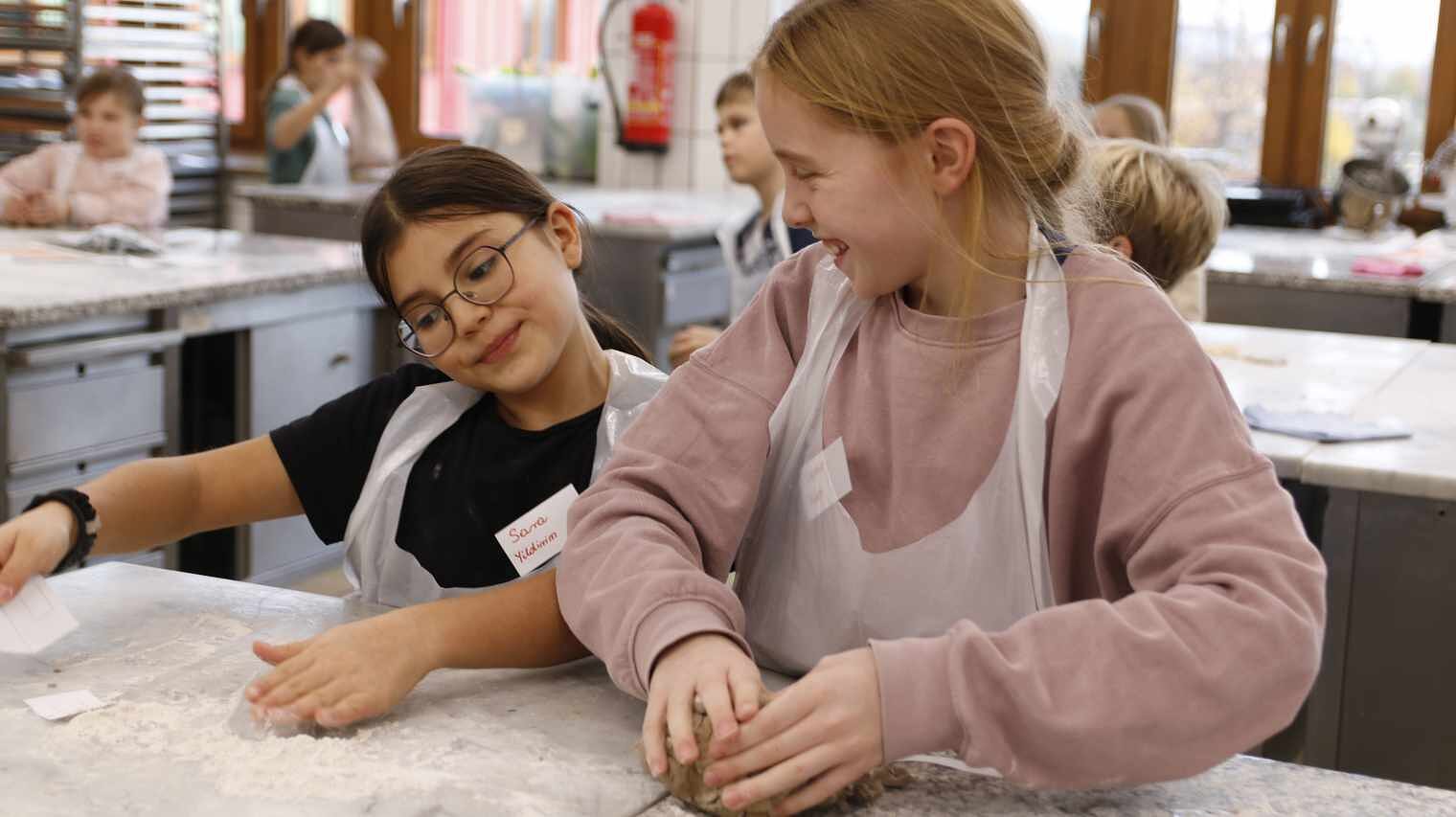 2 Mädchen in einer Backstube, albern mit Mehl herum und kneten Teig zu einer Kugel, beide tragen Schürzen aus Folie und lachen