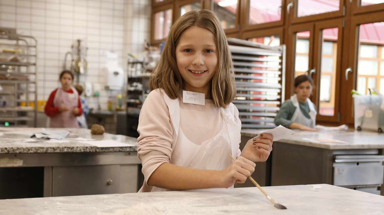 Mädchen in einer Backstube lacht in die Kamera, trägt eine Schürze und pinselt mit einem Pinsel Mehl von der Oberfläche des Tisches