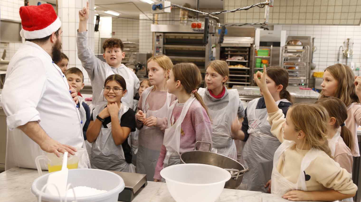 Gruppe von Kindern in der Backstube, links steht ein Bäcker mit Bauch und einer roten Nikolausmütze, Kinder strecken wie in der Schule die Hände hoch