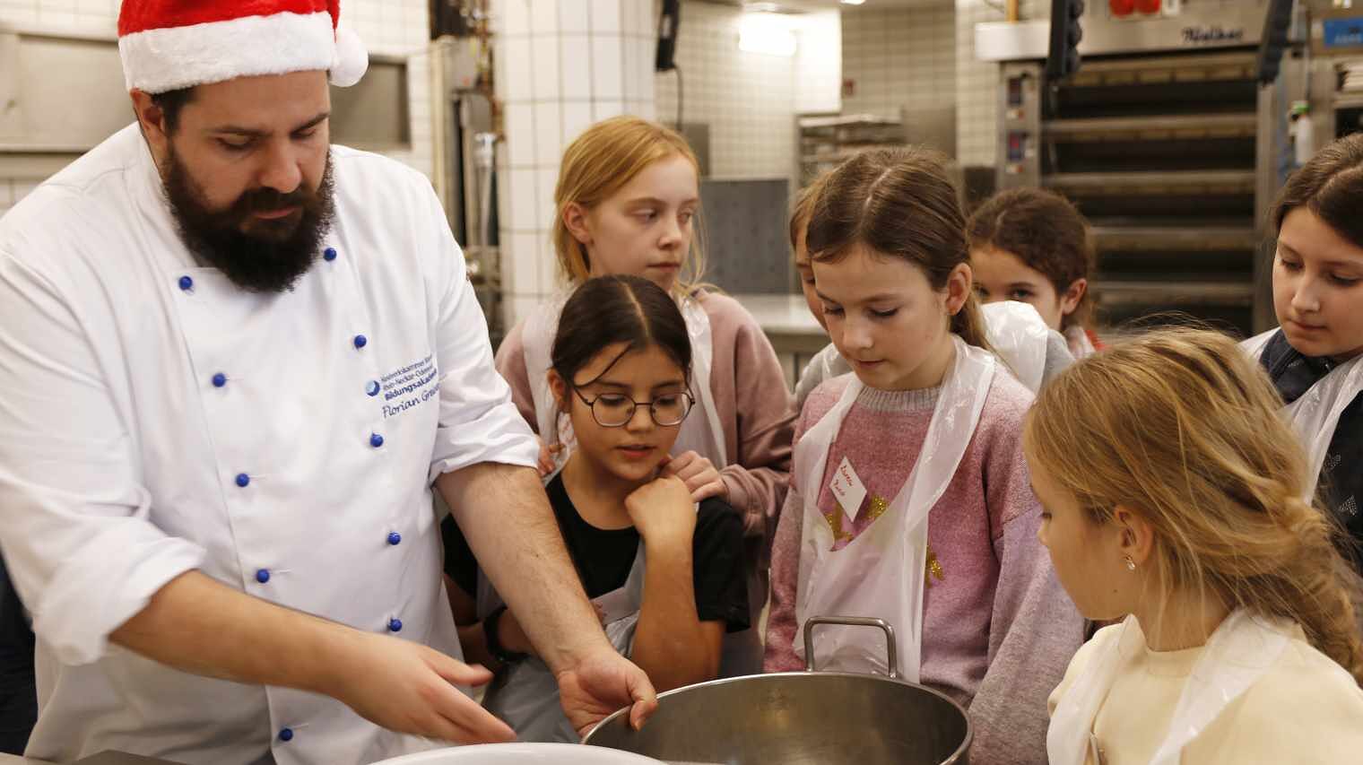 Bäcker am linken Bildrand mit einer roten Nikolausmütze hantiert mit Schüsseln, umringt von Kindern, die ihm gespannt dabei zusehen