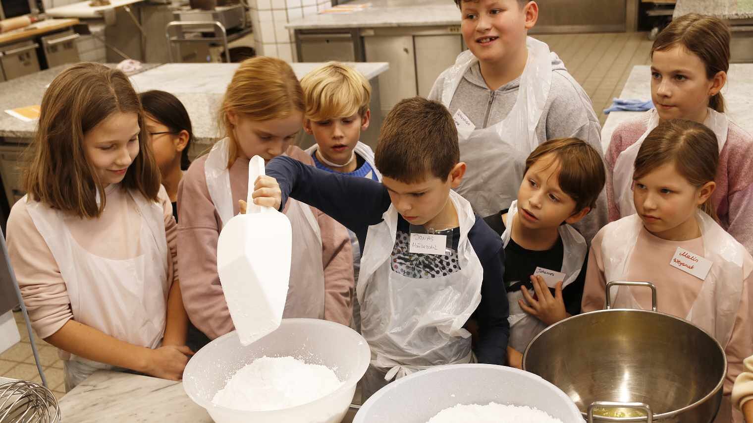 Gruppe von Kindern am Backtisch einer Backstube, der Jung in der Mitte schaufelt mit einer großen Schippe Puderzucker, alle anderen stehen lächelnd um ihn herum