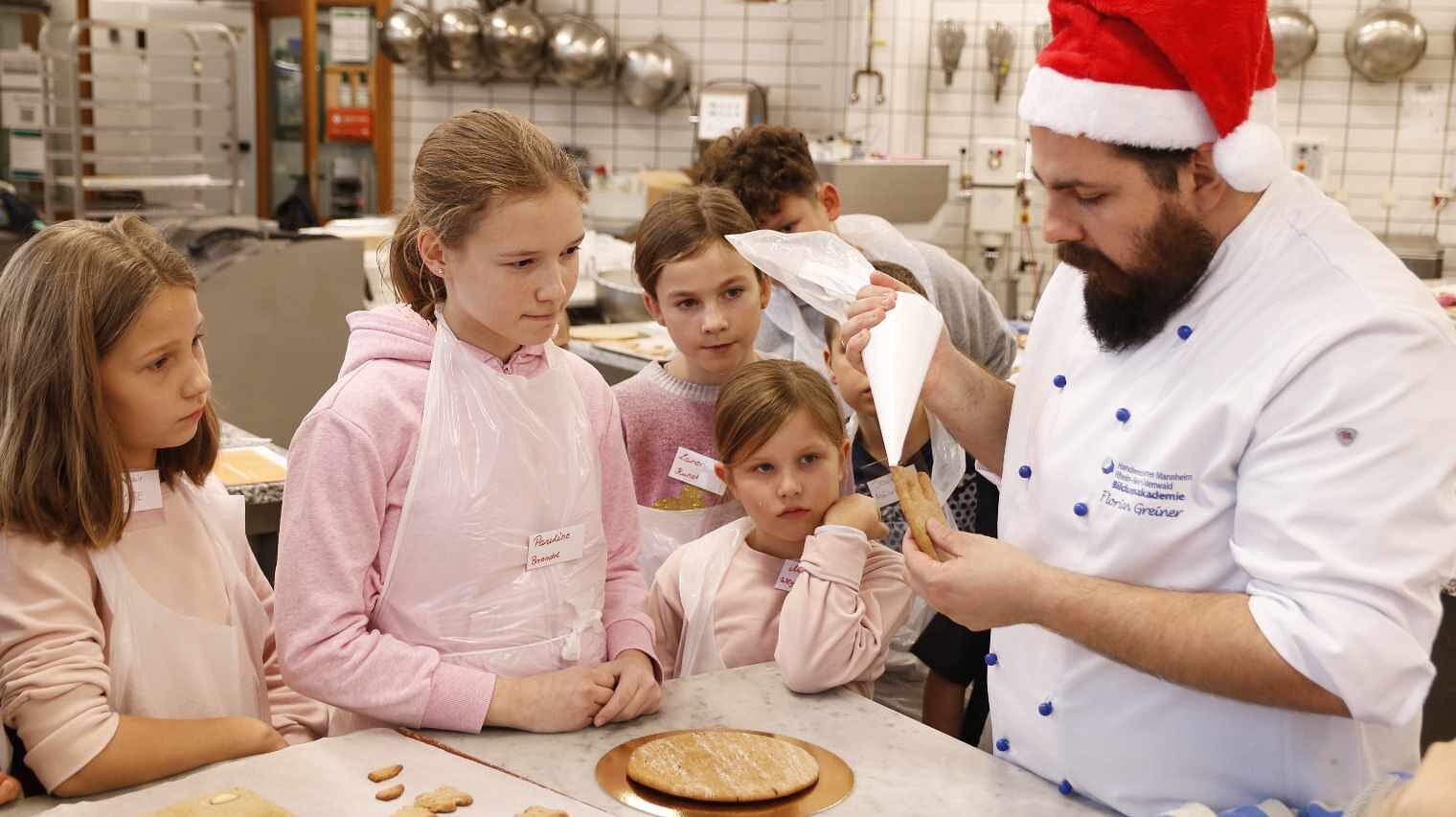 Bäcker in weißer Bäckerkluft mit roter Nikolauszipfelmütze und Spritzbeutel in der Hand, links daneben eine Gruppe Kinder, alle an der Ecke eines Bäckertischs