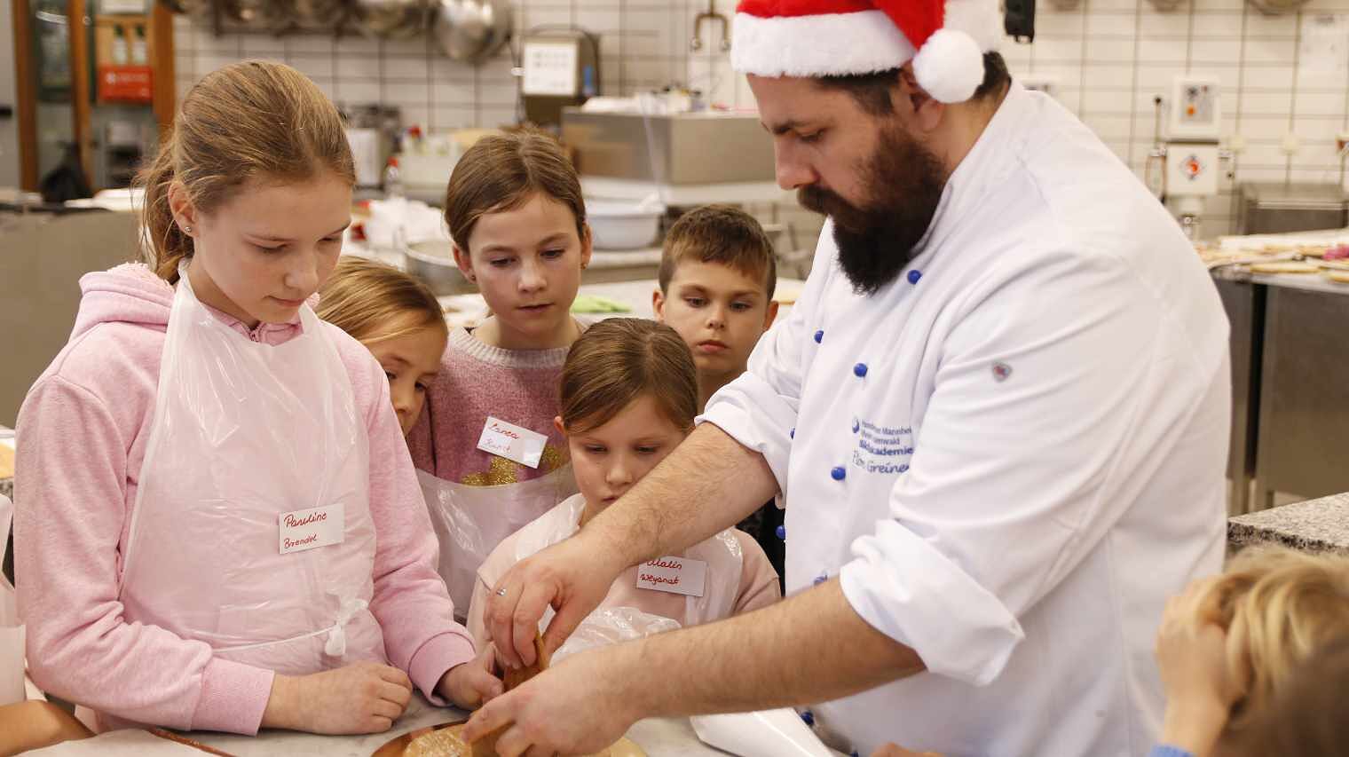 Bäcker mit weißer Kluft, dunklem Vollbart und Nikolauszipfelmütz zeigt einer Gruppe von Kindern etwas an einem Lebkuchenstück