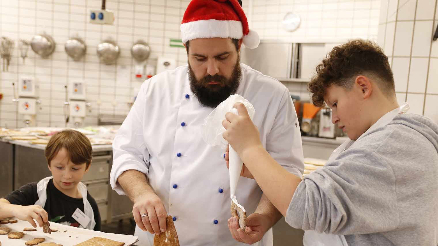 Junge am rechten Bildrand spritzt mit dem Spritzbeutel eine weiße Masse auf die Kante eines Kekses, den ein Bäcker mit Vollbart hält, links ein kleiner Junge