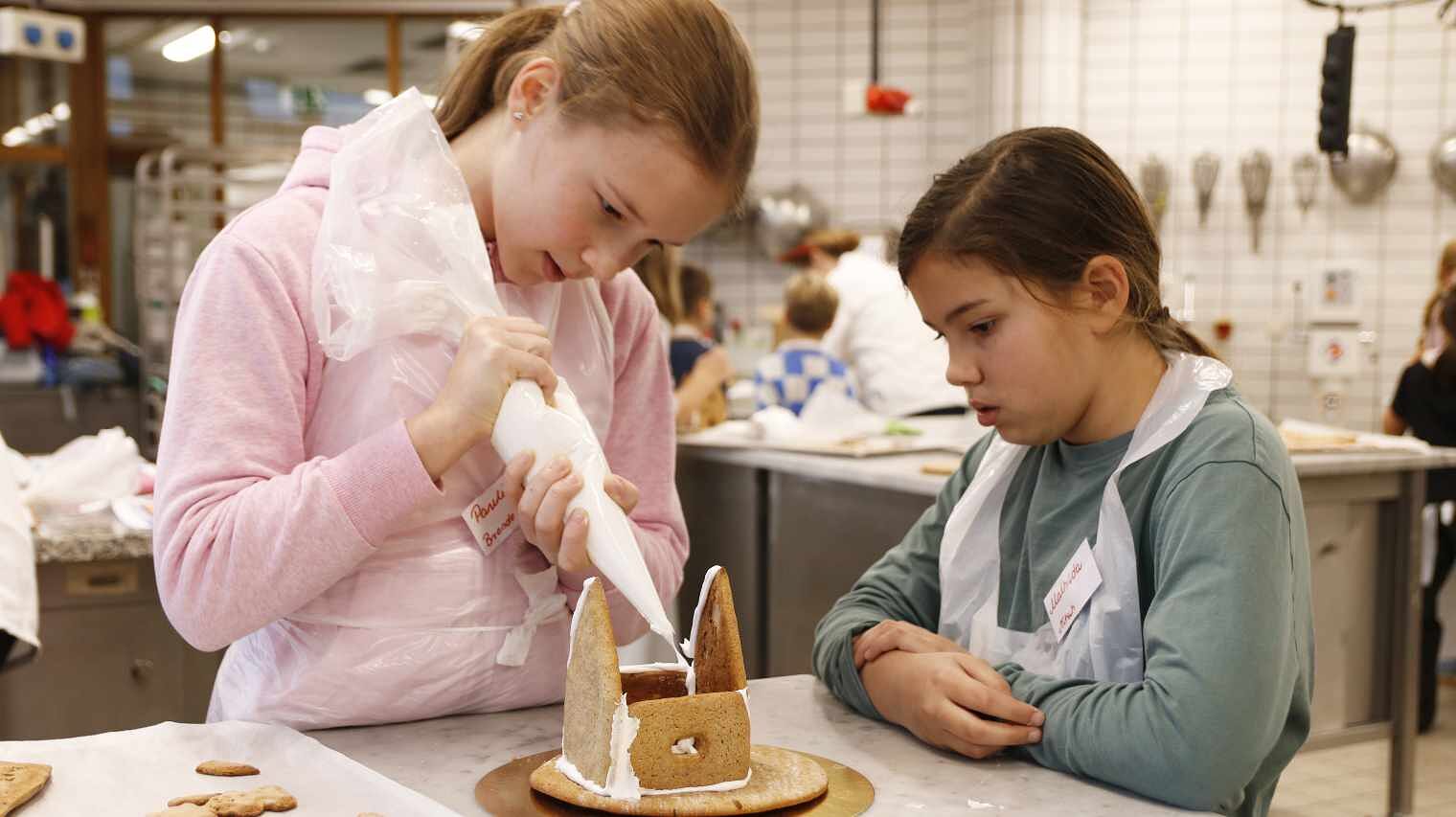 Zwei Mädchen in der Backstube, eine bespritzt mit dem Spitzbeutel die Kanten eines Lebkuchenhauses, die andere schaut am Tisch abgestützt zu