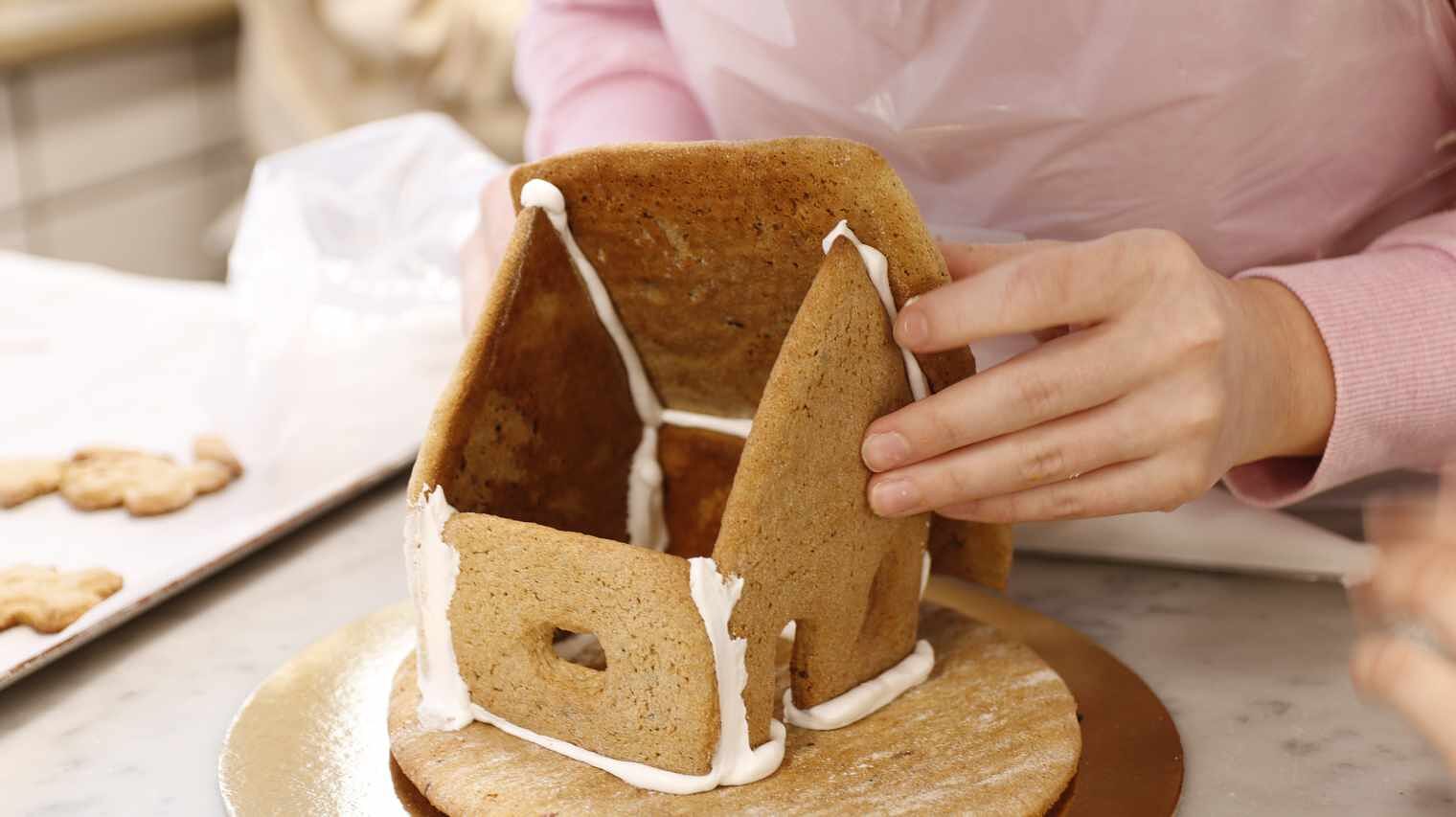 Nahaufnahme eines im Aufbau befindlichen Lebkuchenhauses an dem zwei Hände gerade dabei sind, die hintere Dachhälfte aufzusetzen