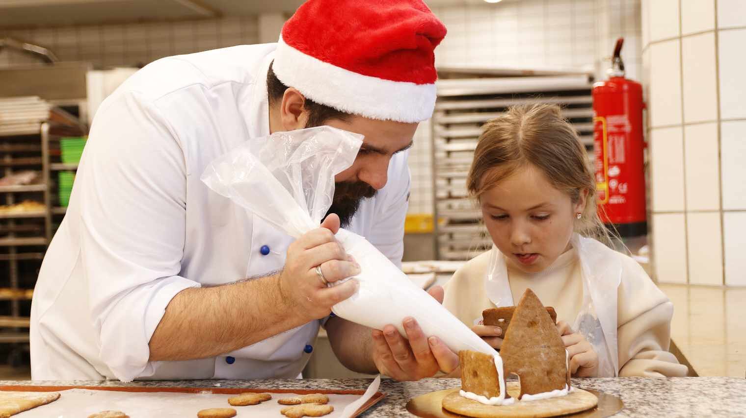 Bäcker mit roter Nikolausmütze und weißer Bäckerjacke spritzt nach vorne gelehnt weiße Masse aus dem Spritzbeutel an die Kanten eines Lebkuchenhauses, ein Kleines Mädchen hält angespannt die Seiten