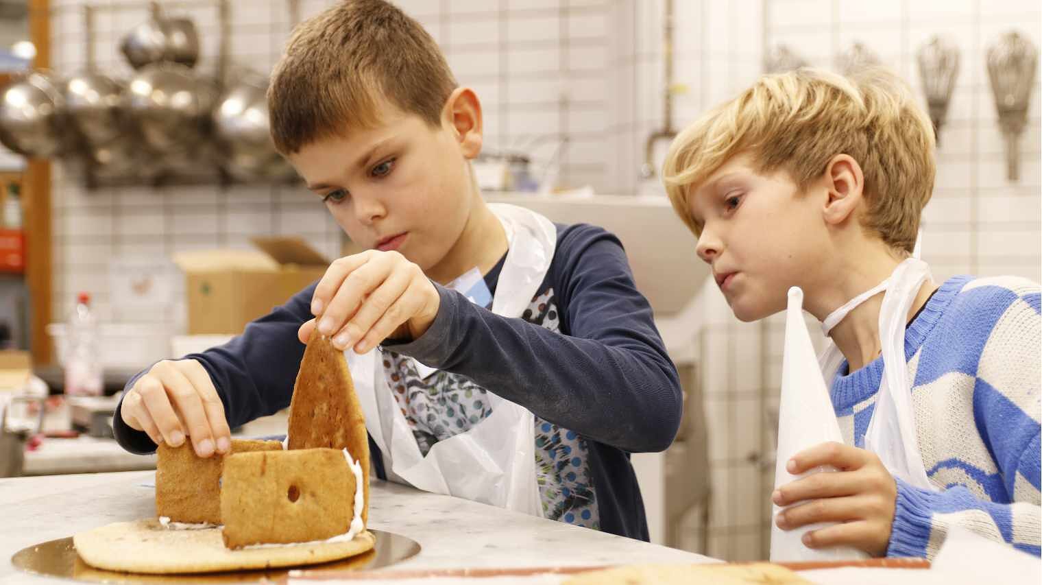 Zwei Jungs bauen ein Lebkuchenhaus zusammen, einer stellt die Vorderwand auf, der andere schaut mit dem Spritzbeutel in der Hand kritisch zu