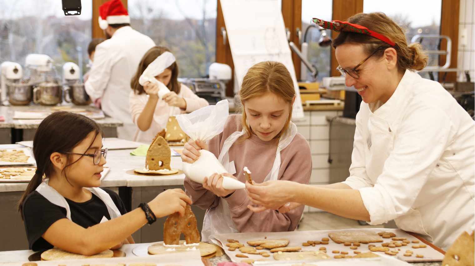 Backstube, in der Kids arbeiten, im Vordergrund zwei Mädchen und eine Bäckerin die gemeinsam über einer mit Gebäcken belegten Fläche und Spitzbeutel arbeiten