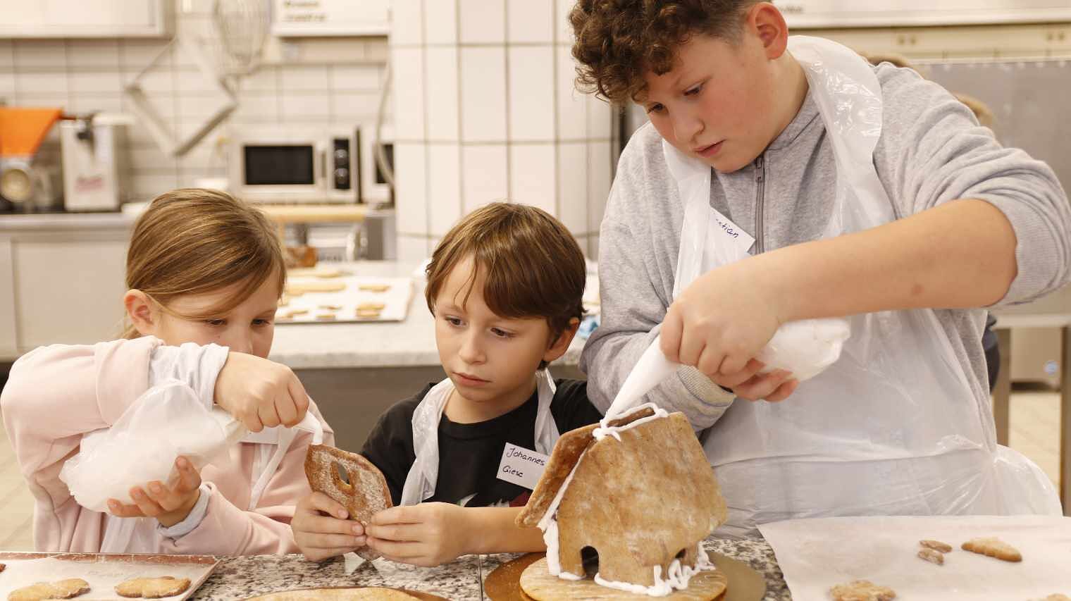 Zwei kleinere Kinder links und ein etwas größerer Junge rechts hantieren mit Spitzbeuteln an Teilen von Lebkuchenhäusern und sich dabei hoch konzentriert