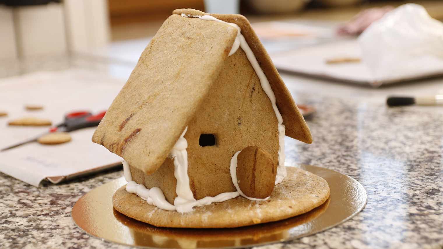 Lebkuchenhaus auf einer Arbeitsplatte, noch nicht fertig, mit offener Keks-Türe und Fensterloch in der Front, weiße Kanten aus Zuckerschaum