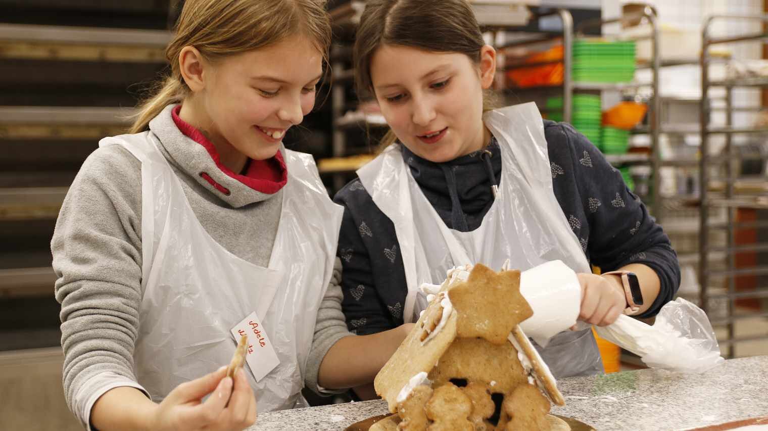Zwei Mädchen in der Backstube, arbeiten gemeinsam an einem Lebkuchenhaus mit dem Spritzbeutel, im Vordergrund das Lebkuchenhaus mit Stern