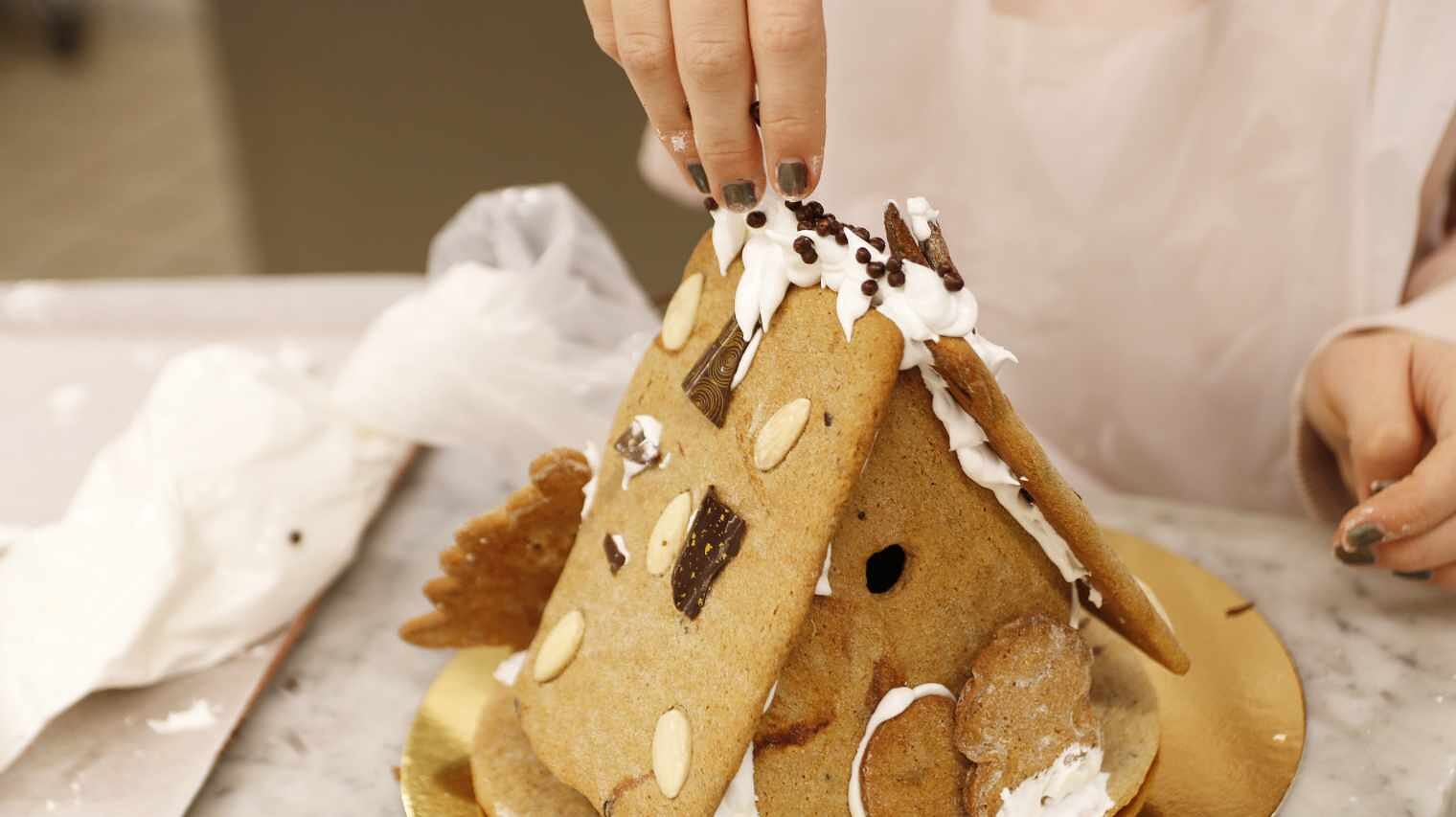 Nahaufnahme eines Lebkuchenhauses mit Mandeln auf dem Dach, oberhalb davon Finger, die Schokostreusel auf den Dachgiebel streuen