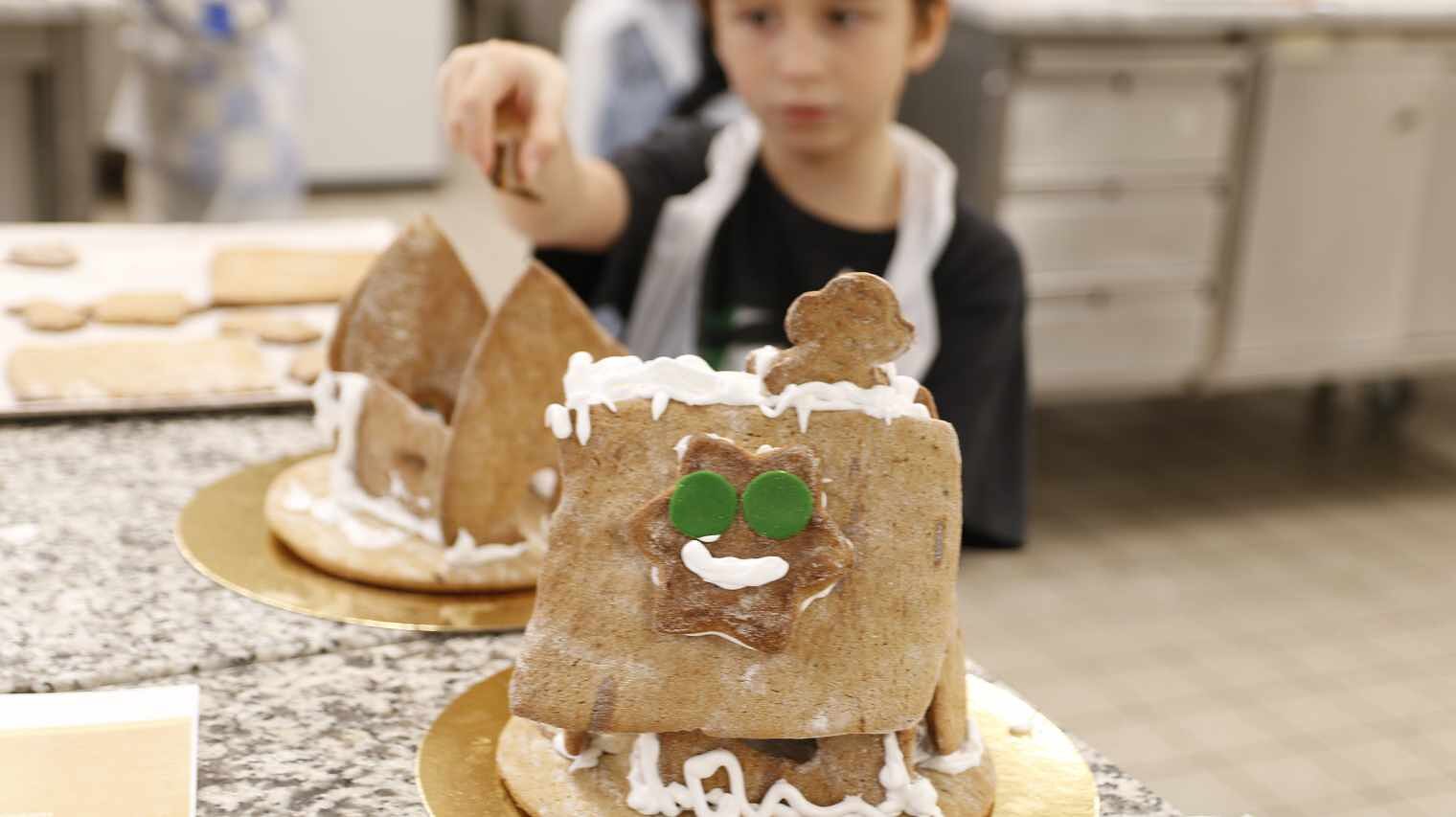 Zwei hintereinander stehende Lebkuchenhäuser in einer Backstube, im Hintergrund verschwommen ein kleiner Junge, der auf eines zeigt