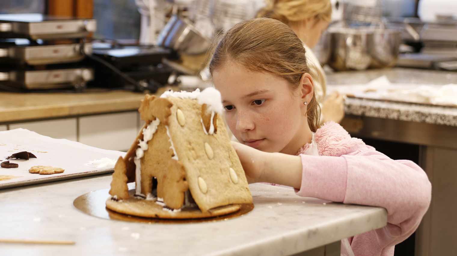 Mädchen mit Zopf und rosa Pulli hockt hinter einer Bäckerplatte, sodass sie auf Augenhöhe mit einem Lebkuchenhaus im Vordergrund ist, an dem sie gerade arbeitet