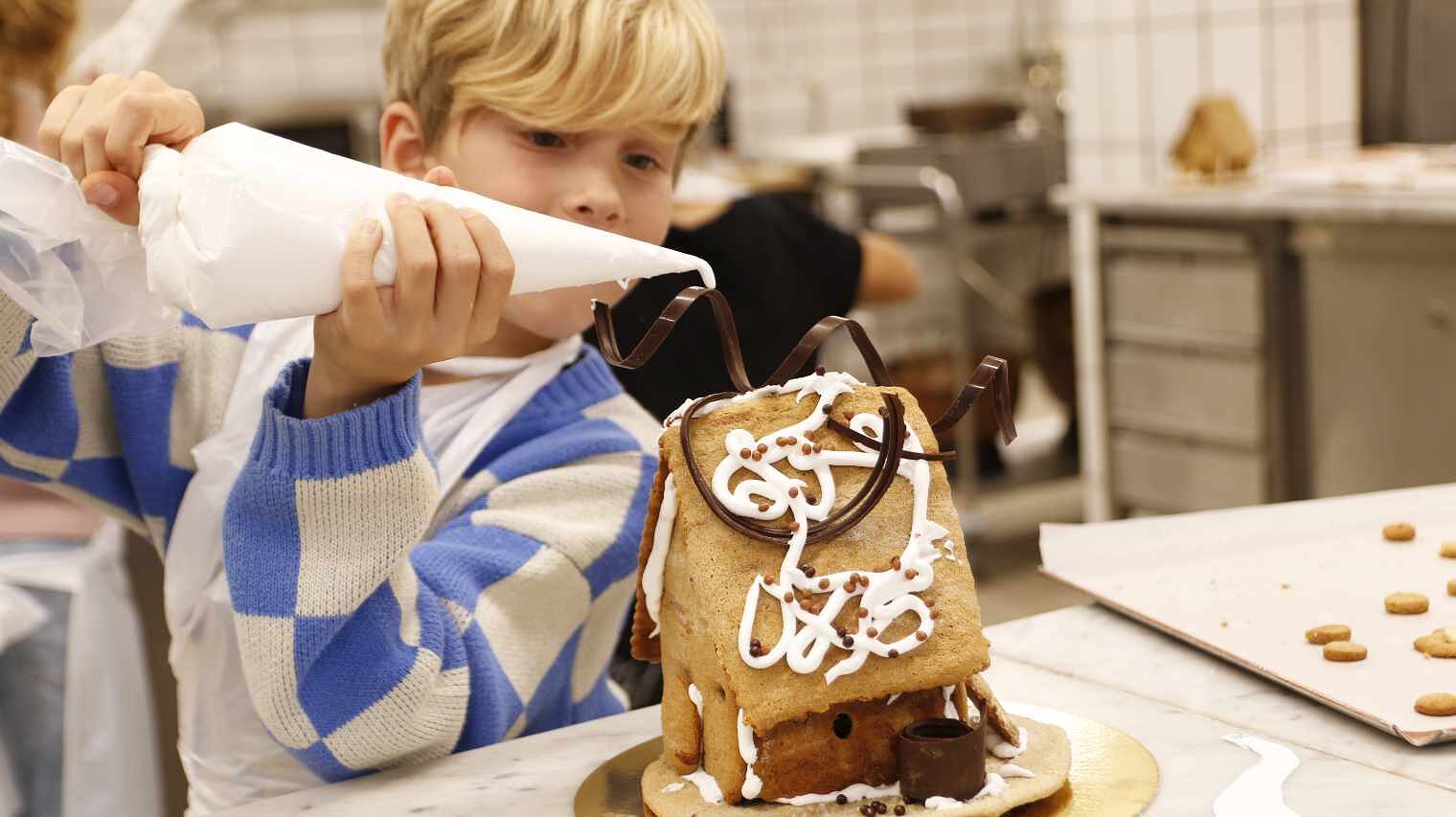 Kleiner blonder Junge mit großkartiertem Pulli hält in einer Backstube einen Spritzbeutel über ein Lebkuchenhaus im Bildvordergrund, um es zu bespritzen
