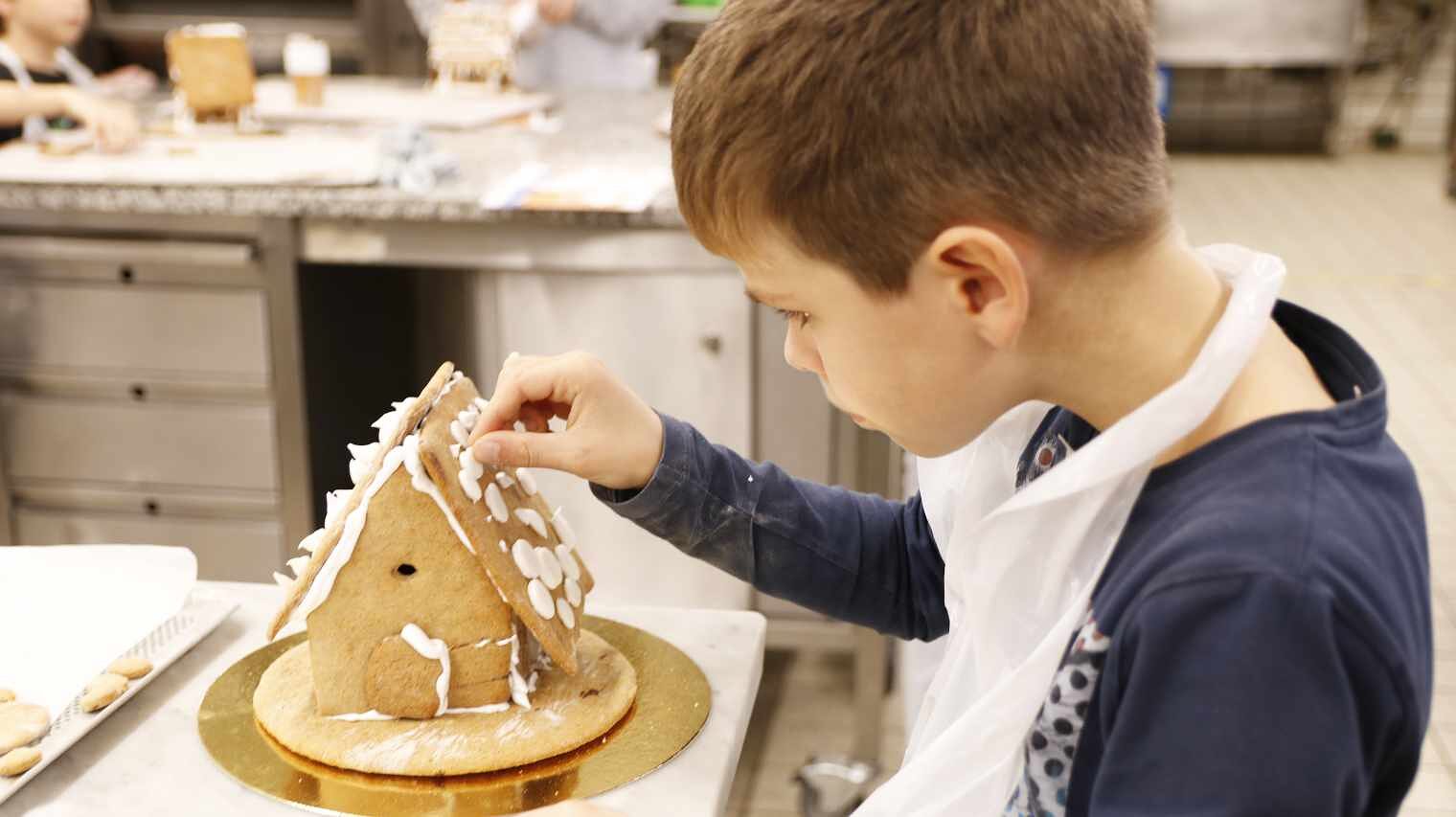 Ausschnitt eines kleinen Jungen in einer Backschürze, der mit vorgebeugtem Kopf auf das Dach eines Lebkuchenhauses Verzierungen klebt