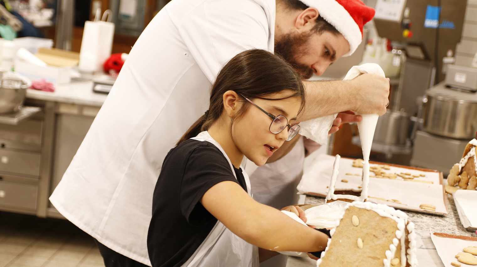 Seitenansicht eines kleinen Mädchens mit dunklem Zopf und Brille an einem Backtisch mit Lebkuchenteilen, dahinter ein Mann im Bäckerkittel mit Zipfelmütze und Spritzbeutel in der Hand