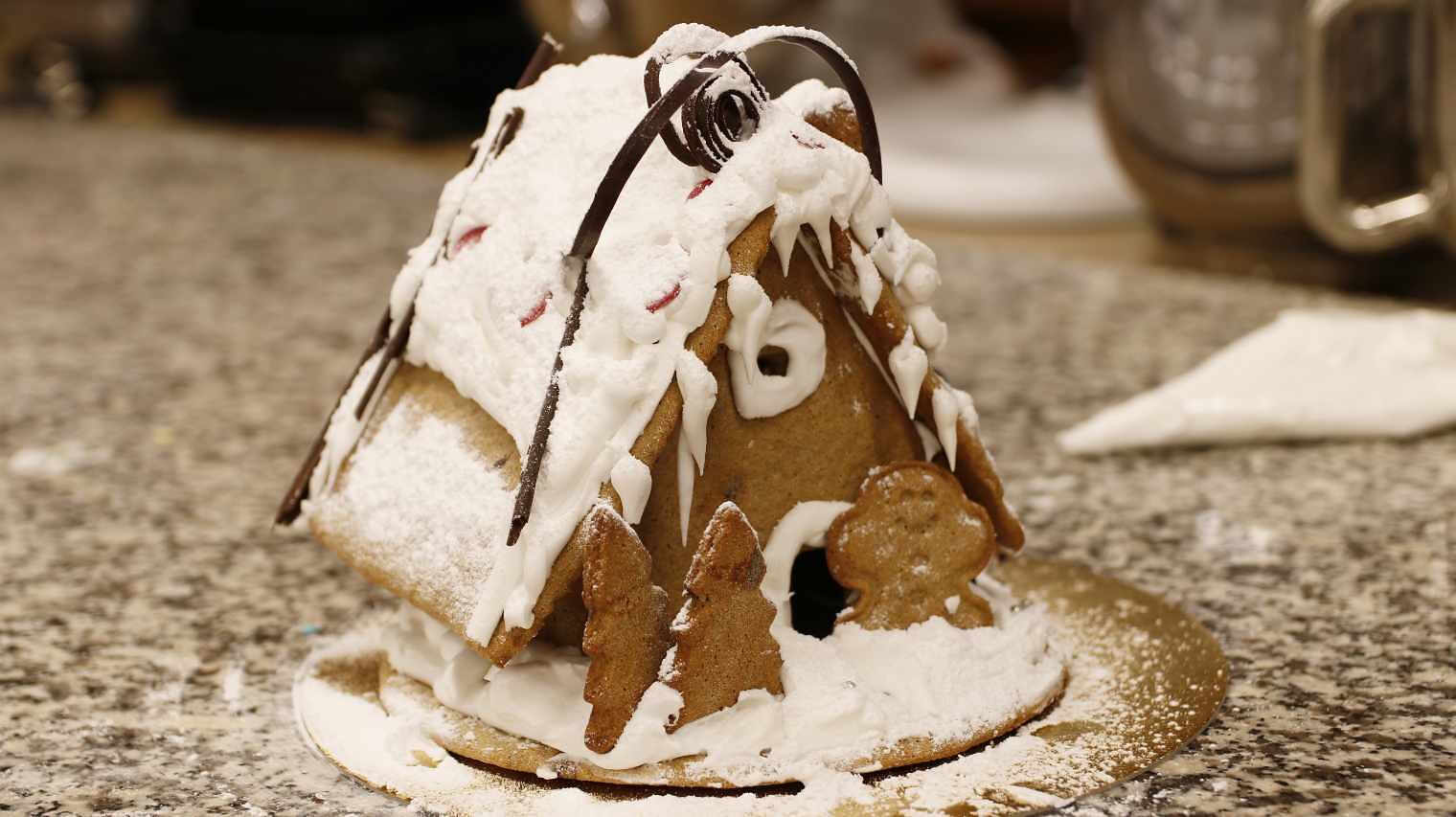 Nahaufnahme eines fertigen Lebkuchenhauses auf einer Steintischplatte mit weiß gezuckertem Dach, Eiszapfen und Bäumen vor der Hütte