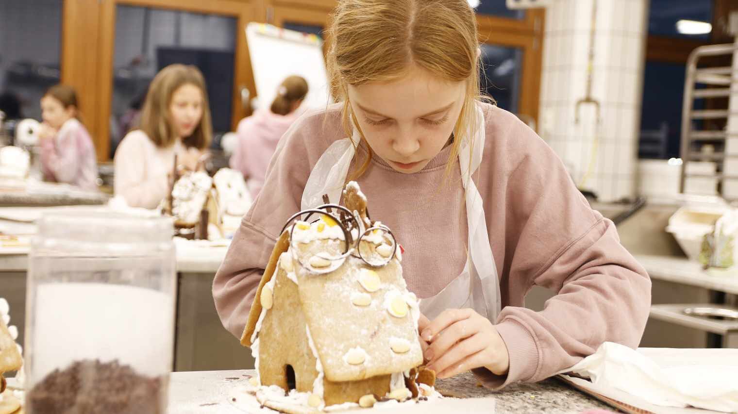 Blondes Mädchen im rosa Pulli in einer Backstube, nach vorne gebeugt über ein Lebkuchenhaus, links vorne ein hohes Glas mit Schokostücken, im Hintergrund verschwommen weitere Kinder