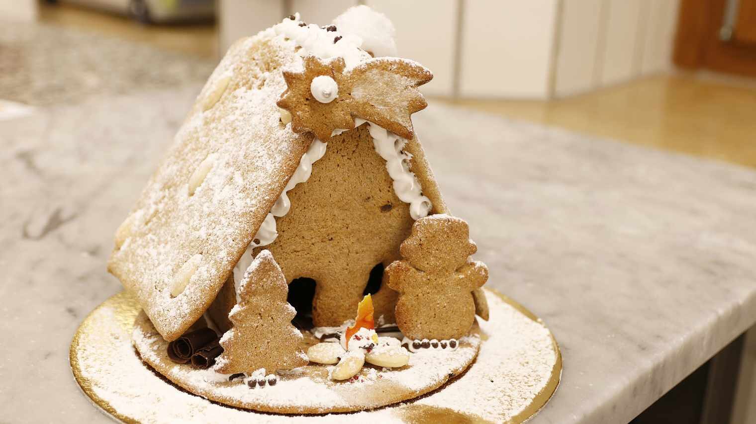 fertiges Lebkuchenhaus auf einer Tischplatte mit gezuckertem Dach, Stern am Giebel, Bäumen und Lagerstelle vor dem Haus