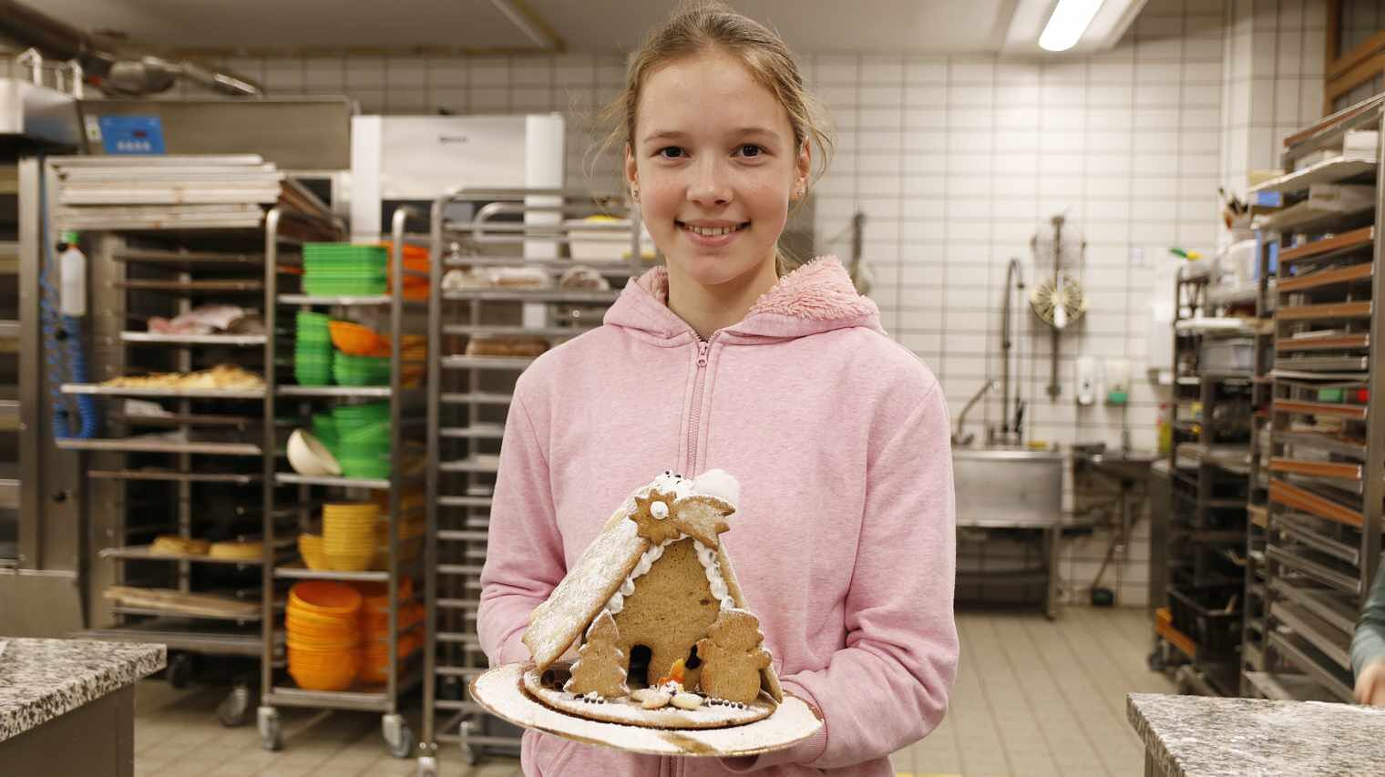 Blondes Mädchen mit Zopf und rosa Kapuzenpulli in einer Backstube, hält lachend ihr fertiges Lebkuchenhaus in die Kamera, im Hintergrund Regale der Backstube