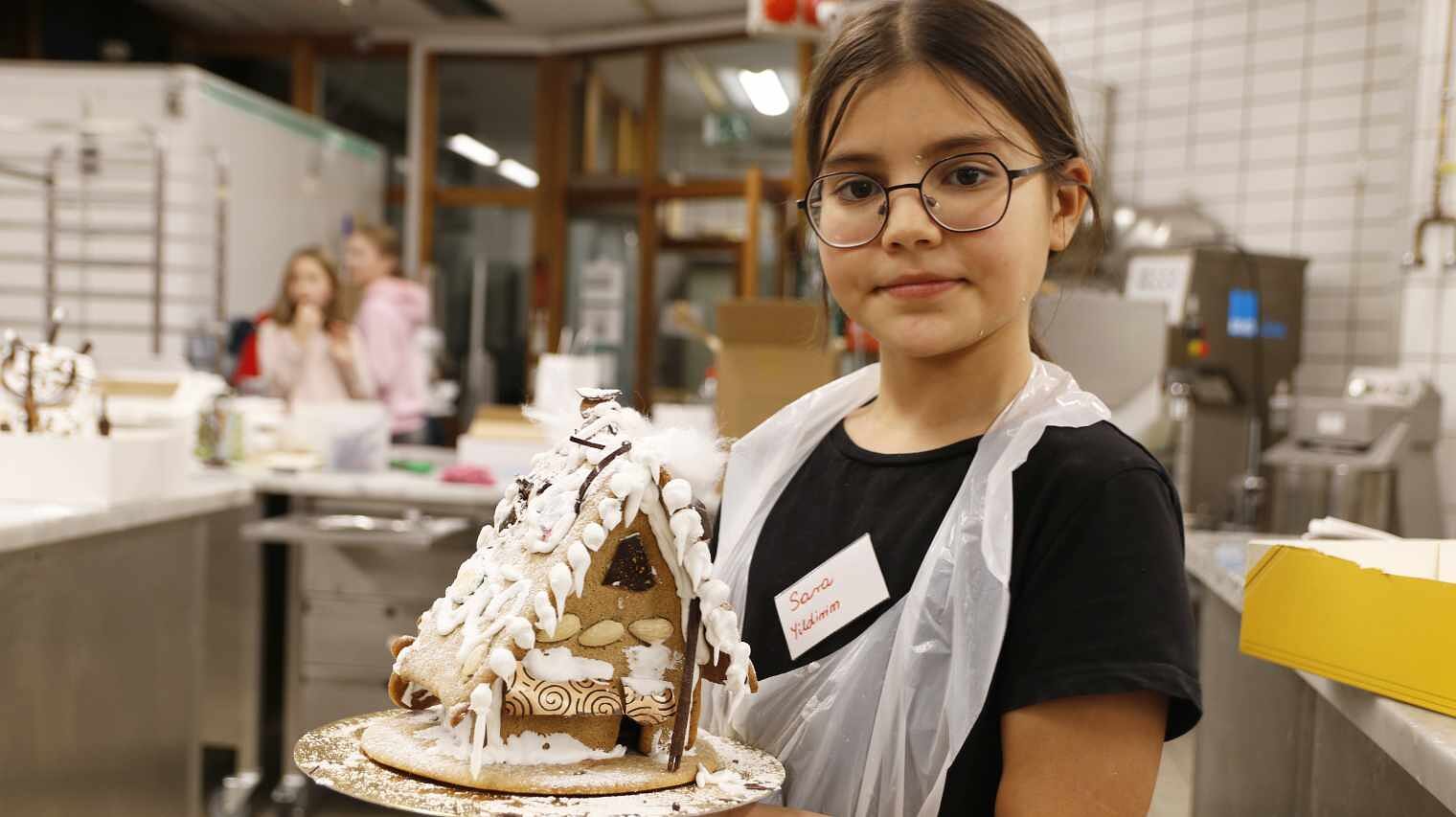Mädchen mit dunklen Haaren und Augen, Brille und Plastikschürze über dem schwarzen T-Shirt zeigt in einer Backstube lächelnd ihr fertiges Lebkuchenhaus in die Kamera