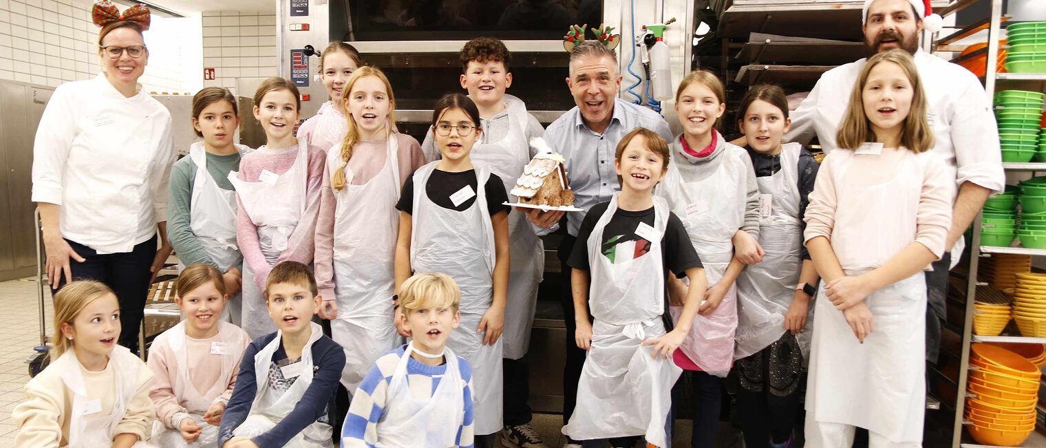 Gruppe von 14 Kindern vor den großen Ofen in einer Backstube, 4 knien vorne links, die anderen stehen, links und rechts flankiert von erwachsenen Bäckern und der Mitte einem Mann, der lachend ein Lebkuchenhaus in die Kamera hält