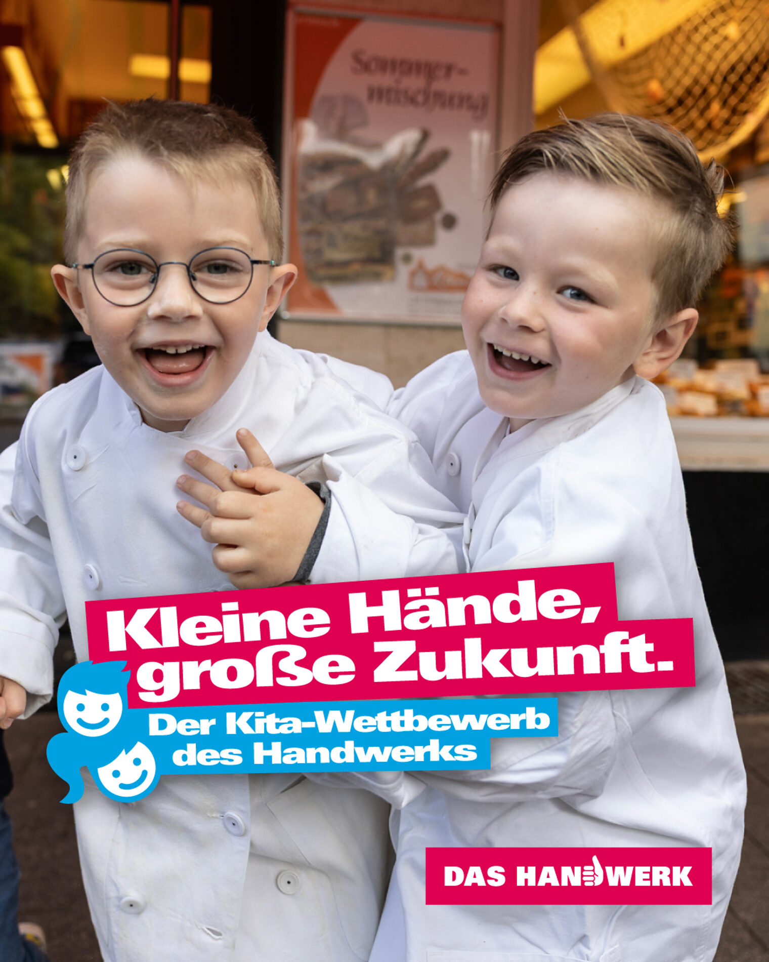 zwei kleine Jungs vor einer Bäckerei tragen viel zu große Bäckerkittel und rangeln lachend miteinander, wobei sie in die Kamera schauen