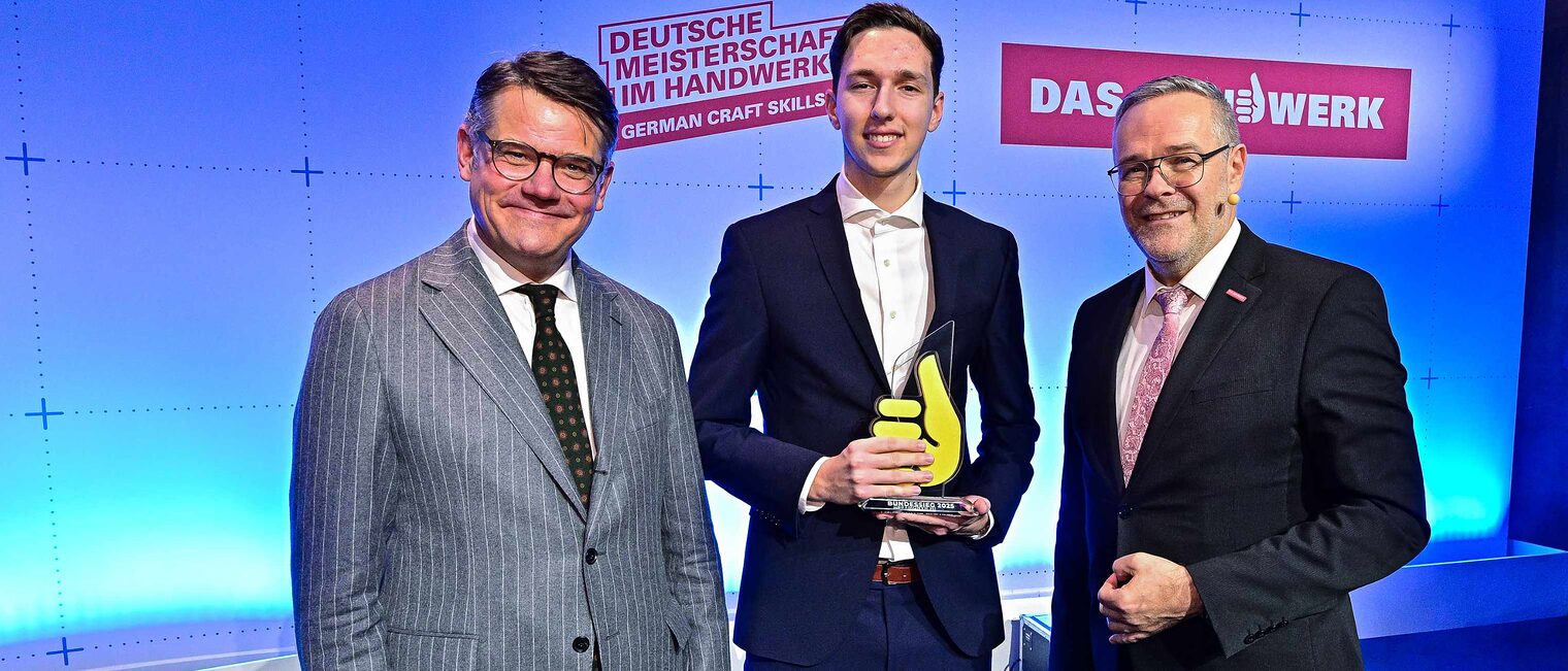 Drei Männer in Anzügen vor einer festlich in Blau beleuchteten Wand mit der Aufschrift "Das Handwerk", der Jüngste in der Mitte hält einen Preis in Form eines Daumens, links und rechts zwei reifere Männer mit Brillen