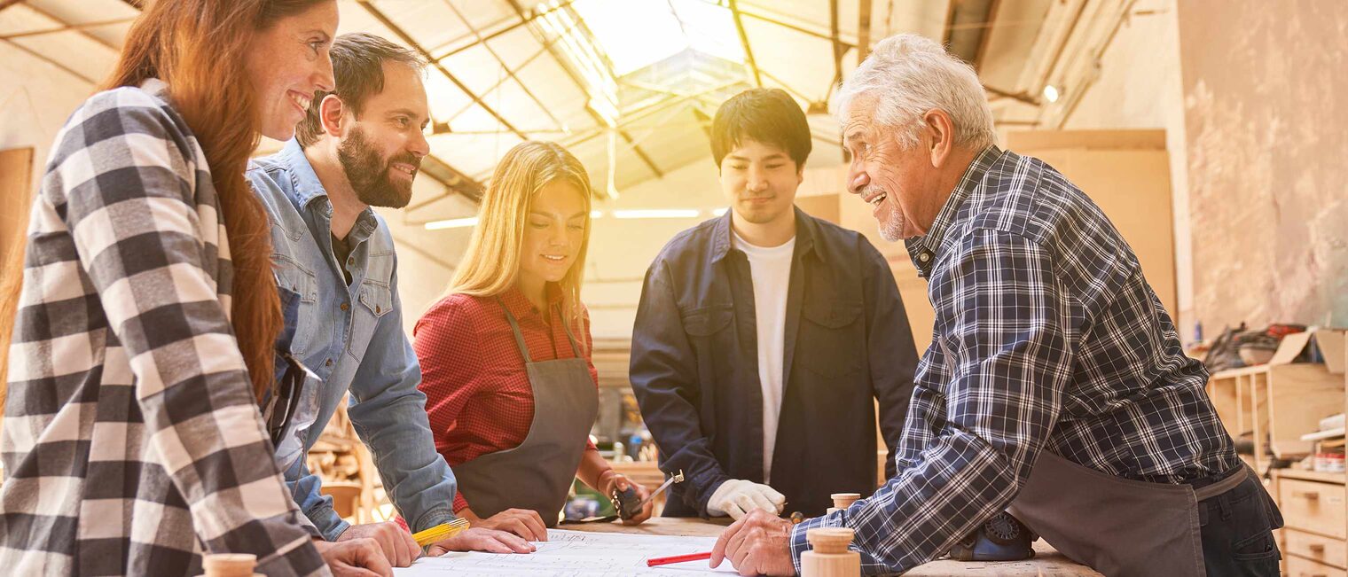 Fünf Menschen stehen in einer Schreinerei rund um einen Tisch, rechts ein alter, weißhaariger Meister mit Schürze zeigt mit dem Stift auf einen Plan, zwei Frauen und zwei Männer sind mit ihm fröhlich im Gespräch