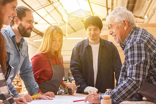 Fünf Menschen stehen in einer Schreinerei rund um einen Tisch, rechts ein alter, weißhaariger Meister mit Schürze zeigt mit dem Stift auf einen Plan, zwei Frauen und zwei Männer sind mit ihm fröhlich im Gespräch