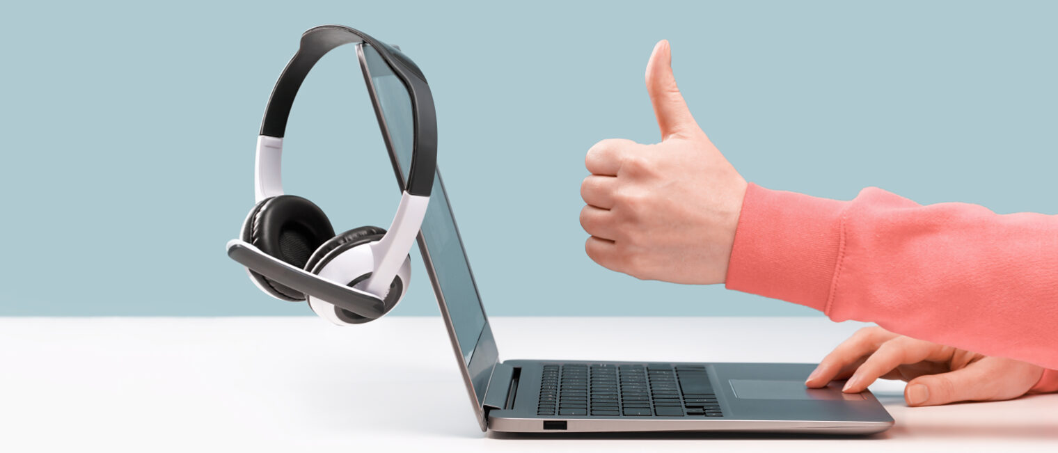 Ein Laptop mit Headphones auf einem weißen Tisch stehende, der von einer Person bedient wird, die den Daumen nach oben zeigt.