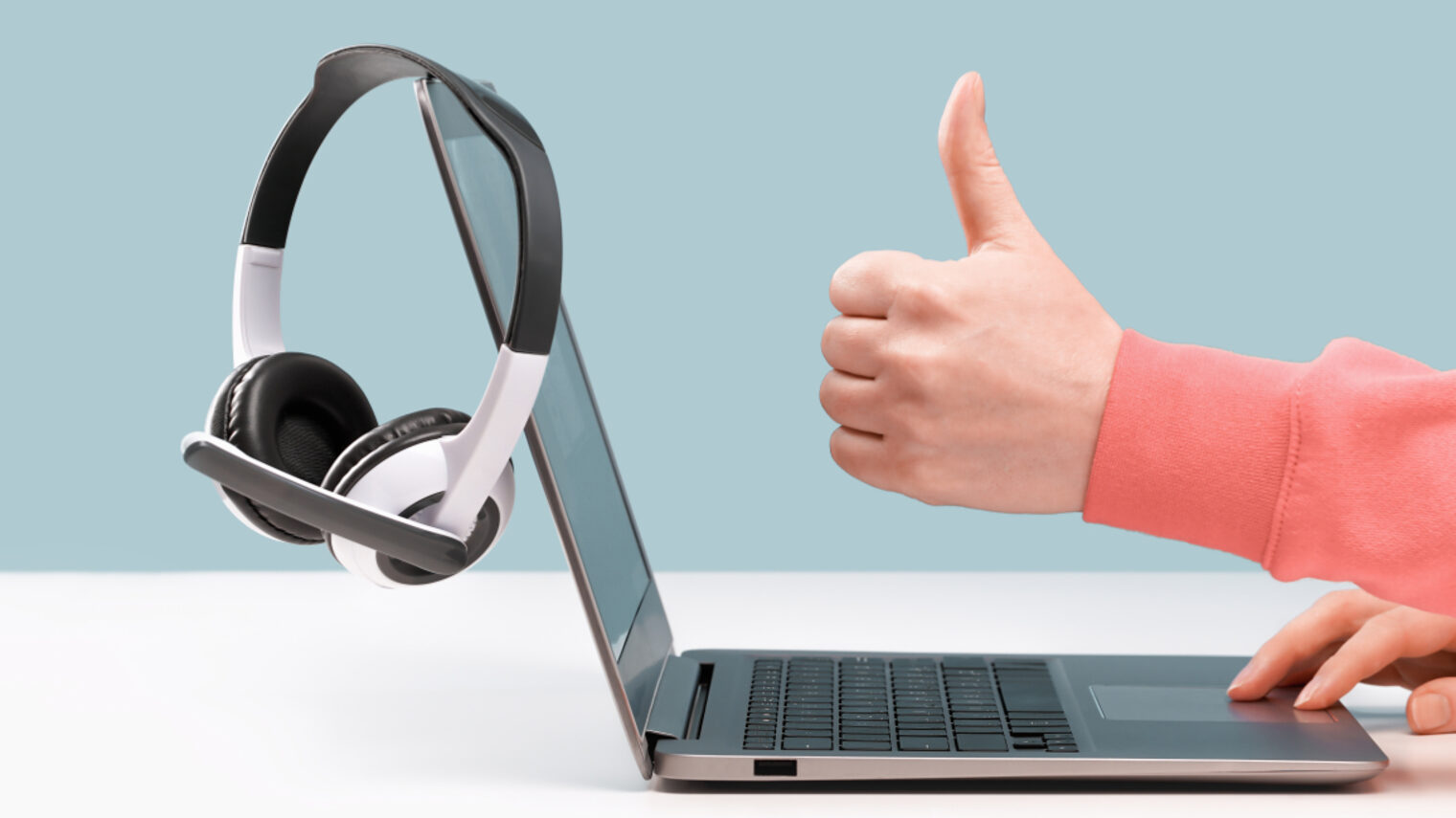Ein Laptop mit Headphones auf einem weißen Tisch stehende, der von einer Person bedient wird, die den Daumen nach oben zeigt.