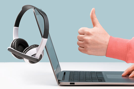 Ein Laptop mit Headphones auf einem weißen Tisch stehende, der von einer Person bedient wird, die den Daumen nach oben zeigt.