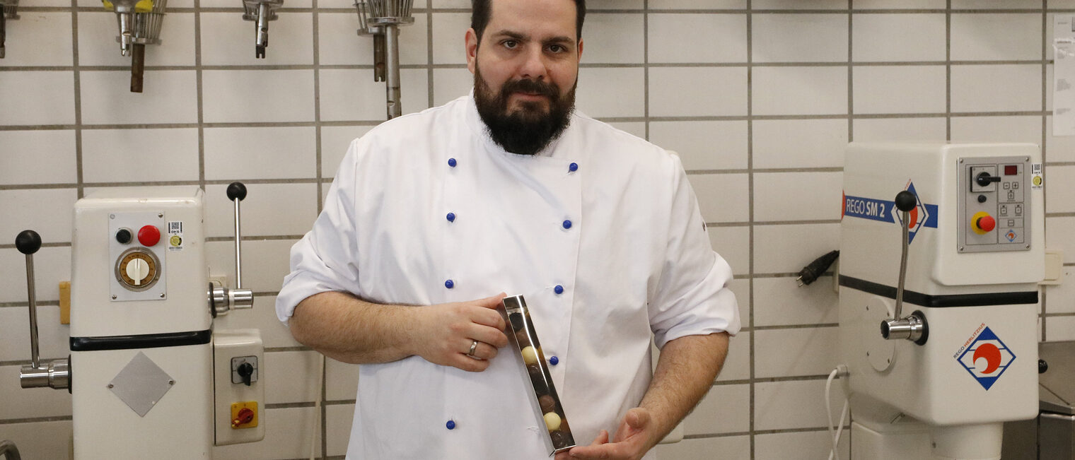 Mann in einer Bäcker-Jacke vor der gekachelten Wand in einer Backstube, links und rechts zwei Geräte zum Backen, hält eine Pralinenschachtel in die Kamera