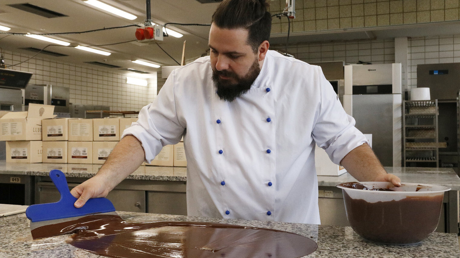 Mann mit dunklem Vollbart und weißer Bäcker-Kleidung streicht mit einem breiten Spatel flüssige Schokolade über die Arbeitsfläche, rechts die Schüssel mit Schoko