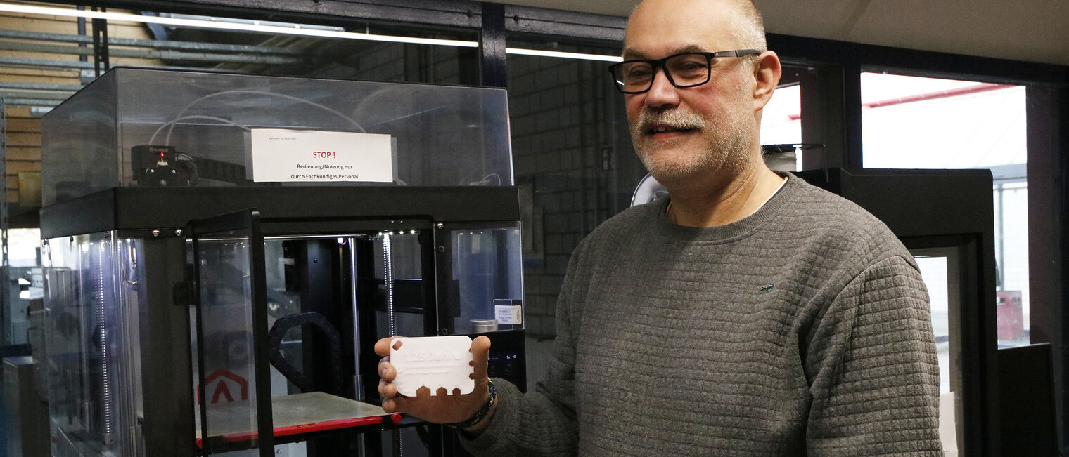 Älterer Mann mit Brille und kurzem weißen Bart steht vor einem 3D-Drucker und hält einen gedruckten flächigen Prototyp in die Kamera
