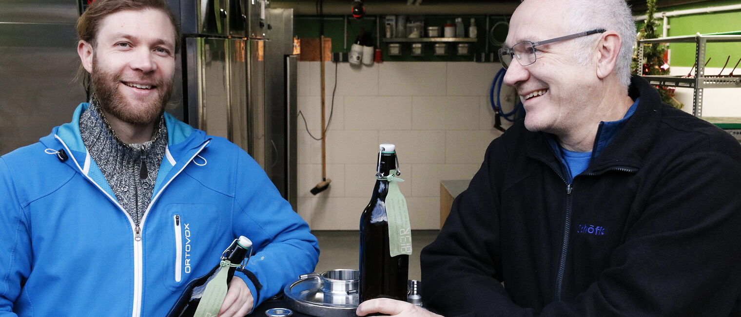 Links ein junger Mann mit roten zum Zopf gebundenem Haar und Vollbart, rechts ein älterer Mann mit weißem Haar und Brille, beide stehen an einem Stehtisch, jeweils eine Bierflasche in der Hand