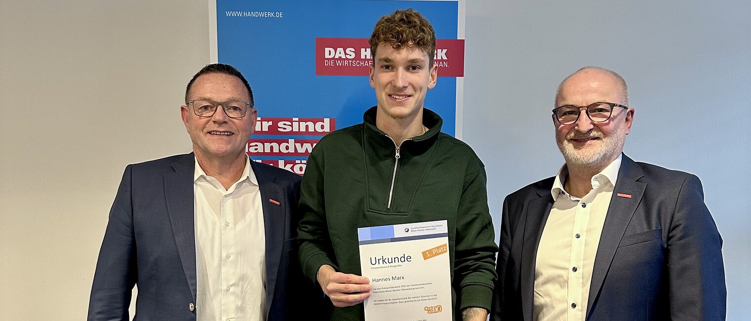 Preisübergabe, drei Männer stehen nebeneinander, der Herr in der Mitte hält sein Zeugnis und seinen Gutschein in die Kamera