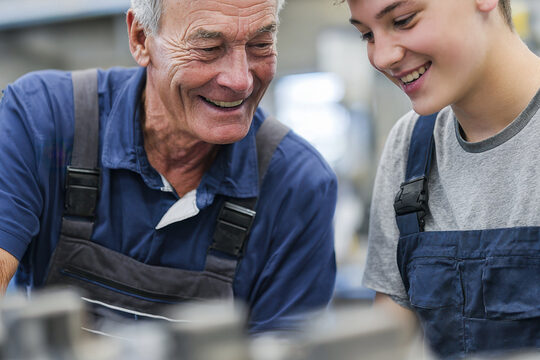 Zwei Handwerker in einer Metallbauwerkstatt, der linke ist ein alter Mann, der als Ausbilder einem jungen Teenager Erklärungen gibt, beide Tragen Latzhosen und lachen