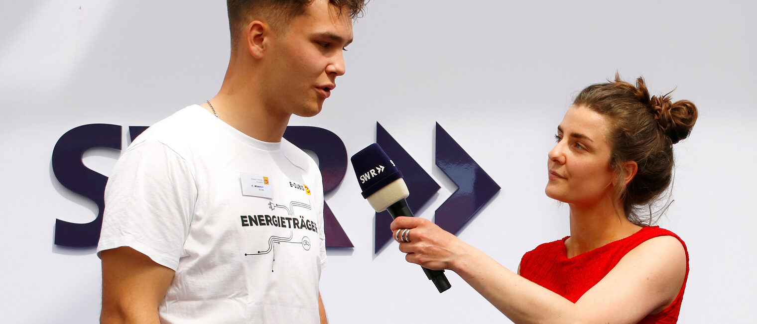 Szene eines Interviews, rechts eine kleine junge Frau mit rotem Top und locker zum Knoten hochgesteckten braunen Haaren, die dem jungen, großen Mann links ein Mikrophon entgegenstreckt, im Hintergrund verdeckt die Buchstaben SWR