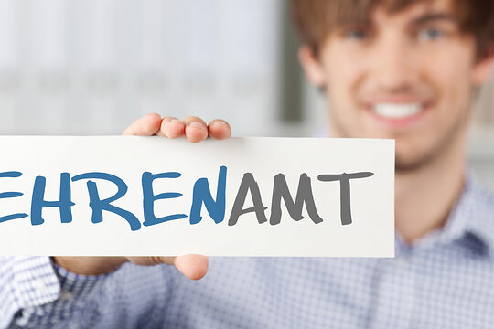 Junger Mann mit in die blauen Augen gekämmtem kurzem Haar hält ein Schild mit der Aufschrift "Ehrenamt" in die Kamera, Gesicht und Körper verschwimmen im Hintergrund