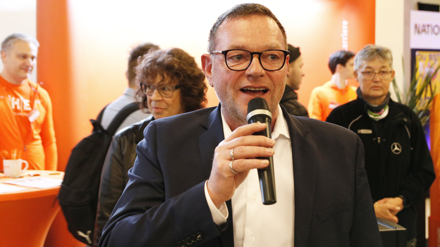 Mann mit Brille und Anzug hält ein Mikrophon in der Hand und spricht hinein, im HIntergrund ein Messeambiente mit Personen
